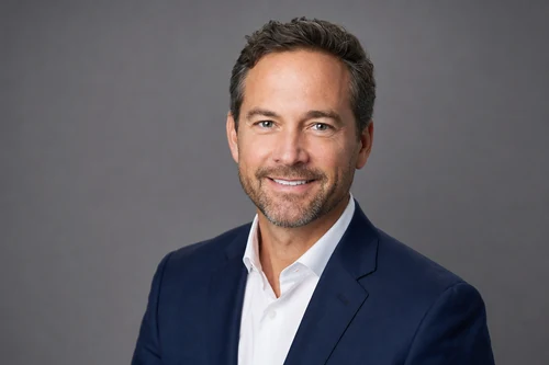 Um headshot profissional estilo LinkedIn de um homem de terno azul-marinho contra um fundo cinza neutro com iluminação de estúdio suave e uniforme.